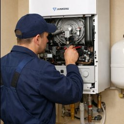 Instalator w niebieskim kombinezonie serwisuje wiszący piec gazowy Junkers, używając narzędzi. Widoczne rury i elementy instalacji. Jasne wnętrze.