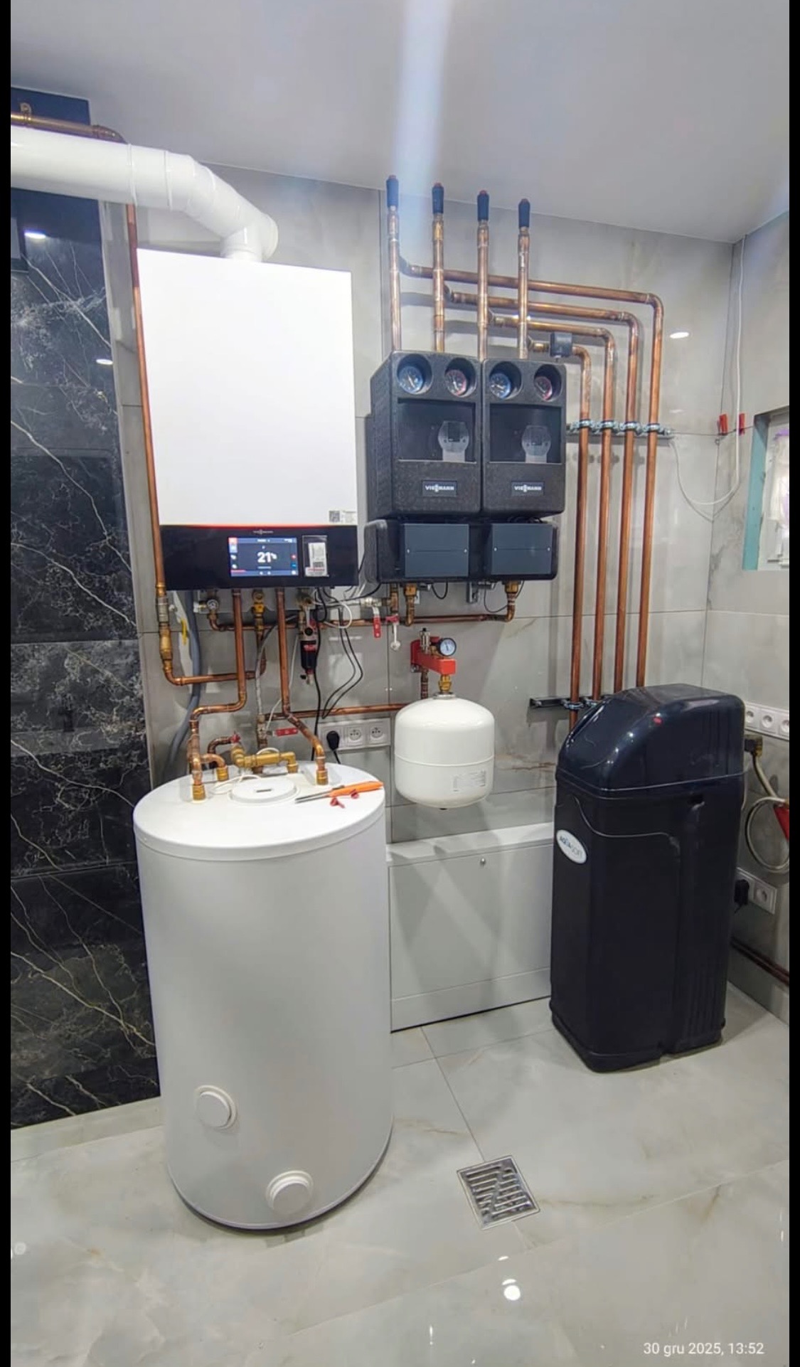 Nowoczesna kotłownia z białym zasobnikiem, piecem gazowym Viessmann i rozbudowaną instalacją miedzianą na tle marmurowej ściany i płytek podłogowych.