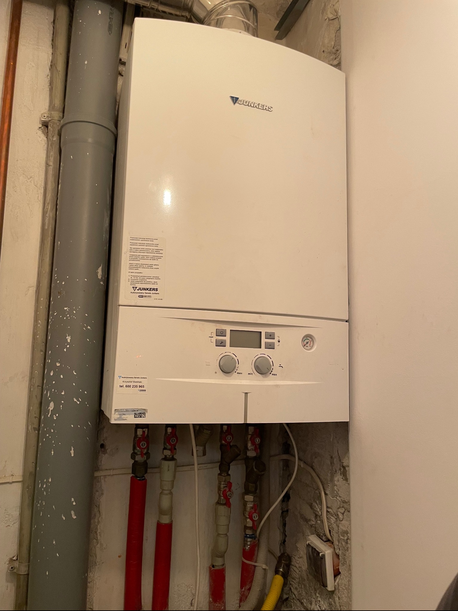 Biały piec gazowy Junkers zamontowany na ścianie z widocznymi rurami doprowadzającymi gaz i wodę. Szare rury odprowadzające spaliny. Ściana w surowym stanie.