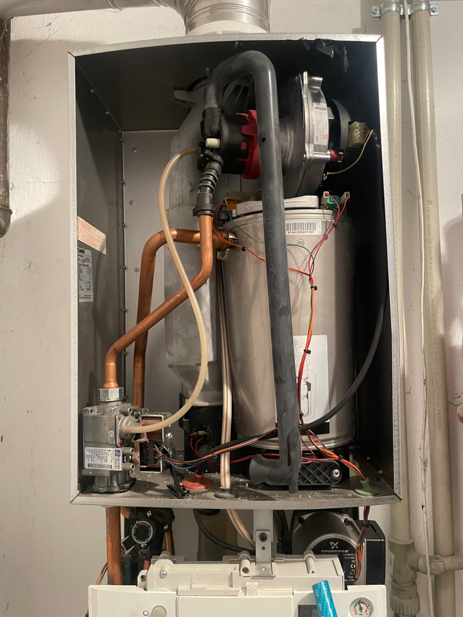 Otwarty piec gazowy z widocznymi elementami instalacji: rurami miedzianymi, przewodami elektrycznymi i pompą Grundfos. Widoczne detale montażu i podłączenia.