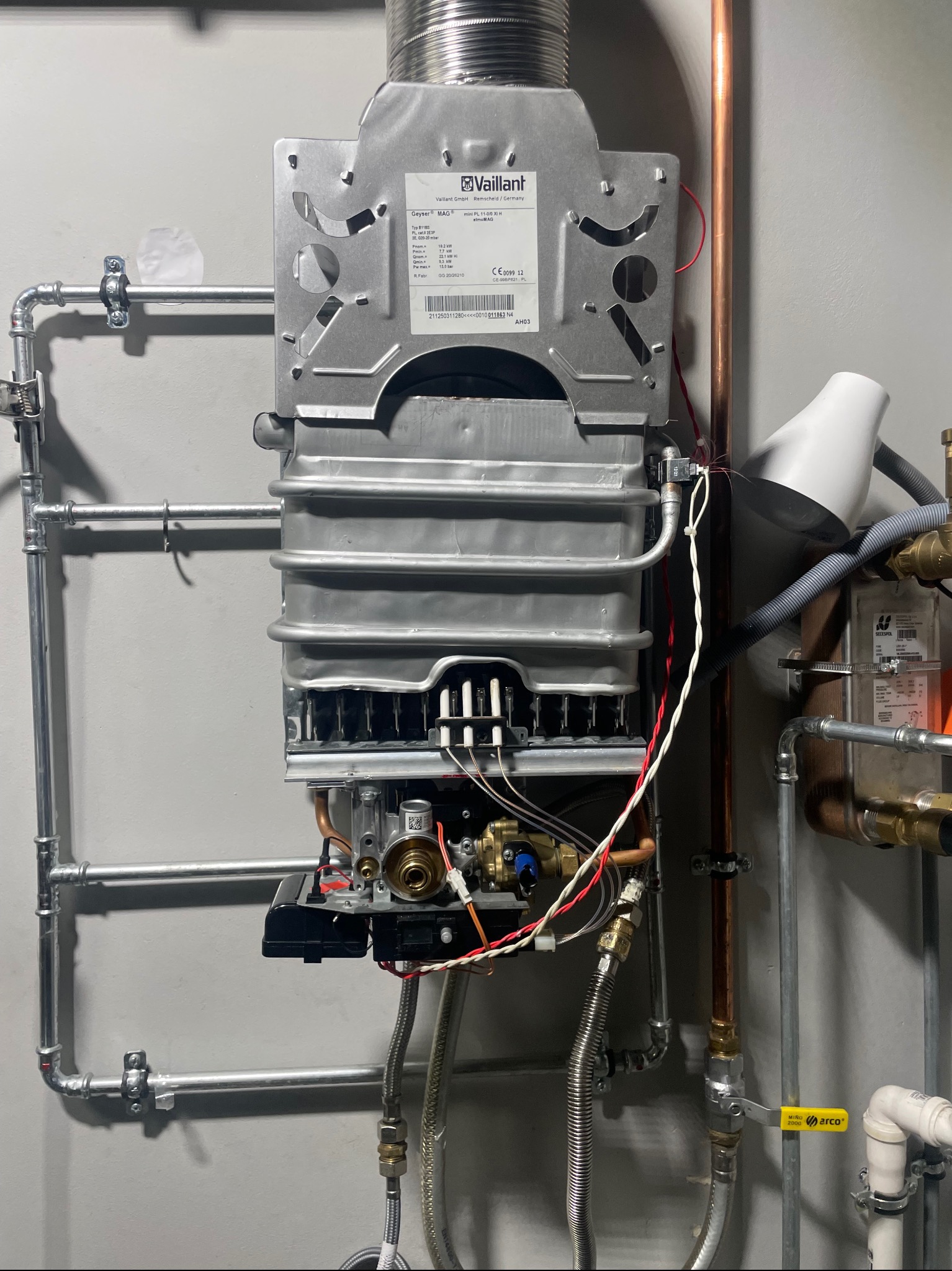 Nowoczesny, wiszący piec gazowy Vaillant z podłączonymi rurami miedzianymi i instalacją, zamontowany na szarej ścianie. Widoczne detale przyłączy i komponentów.