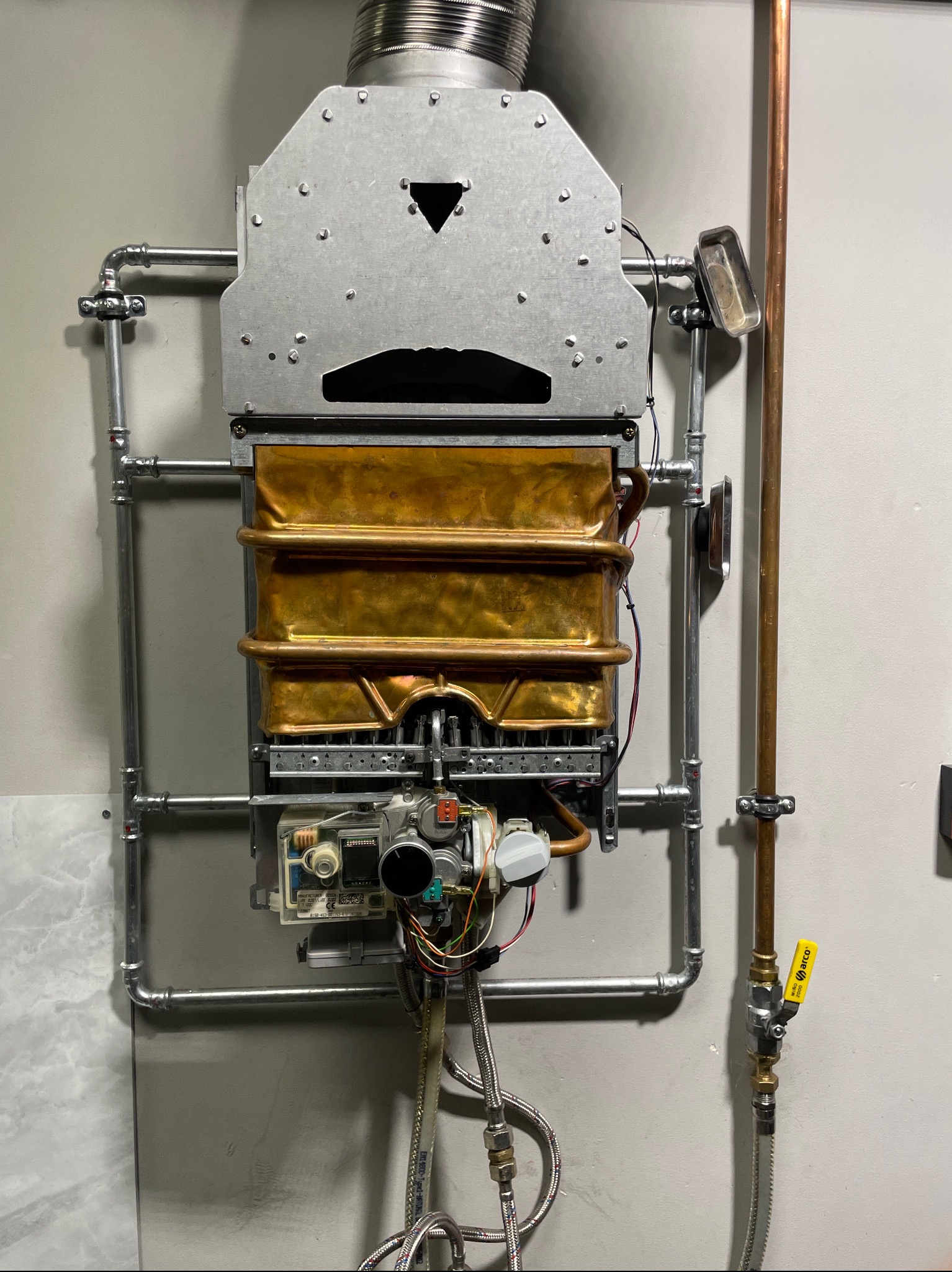 Nowoczesny, wiszący piec gazowy z miedzianym wymiennikiem ciepła i metalową obudową, zamontowany na szarej ścianie z widocznymi rurami doprowadzającymi gaz.