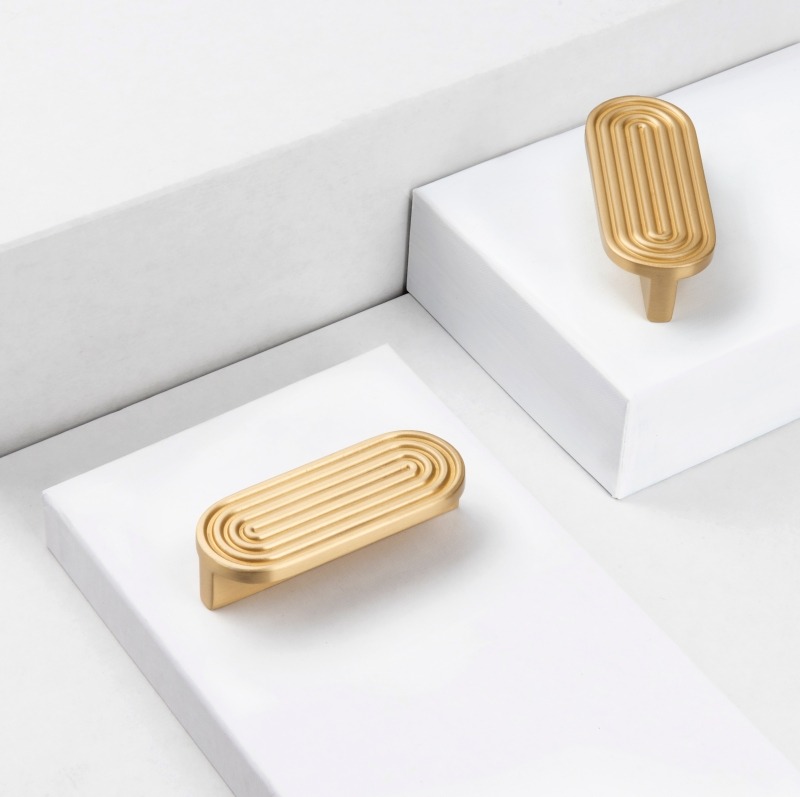 Dwie złote, owalne gałki meblowe z prążkowanym wzorem na białym tle. Jedna leży, druga stoi, ukazując detale i minimalistyczny design.