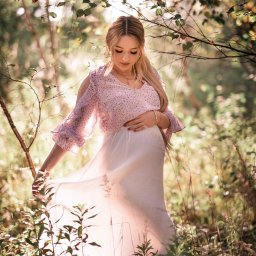 Dagmara Pużyńska Fotografia - Kobieta w ciąży w zwiewnej sukience, delikatnie dotyka brzucha, pozując w leśnej scenerii z promieniami słońca przebijającymi przez gałęzie. Romantyczna atmosfera.