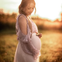 Dagmara Pużyńska Fotografia - Kobieta w ciąży w zwiewnej sukience, trzymająca mały model samochodu na brzuchu, w ciepłym świetle zachodzącego słońca na łące. Romantyczna sesja ciążowa.