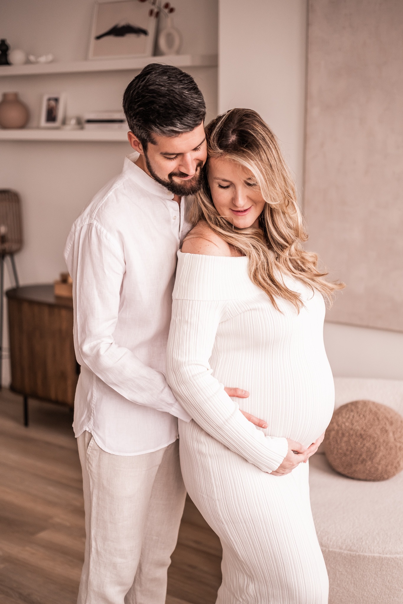 Uśmiechnięty mężczyzna obejmuje ciężarną kobietę w białej sukience, oboje patrzą na jej brzuch. Jasne, minimalistyczne wnętrze w tle.