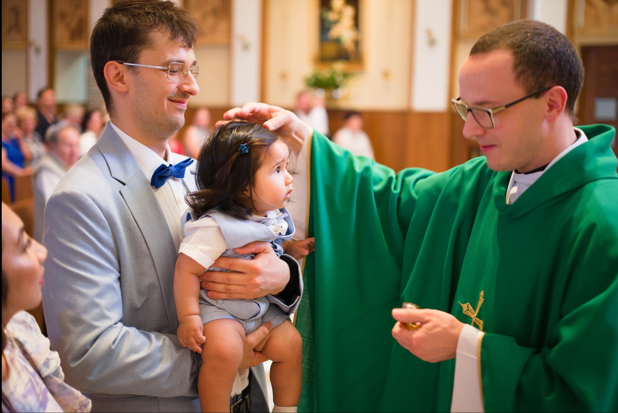 Uśmiechnięty ojciec trzyma dziecko podczas chrztu, ksiądz w zielonej szacie błogosławi. W tle widoczni goście, wnętrze kościoła, ciepłe oświetlenie.