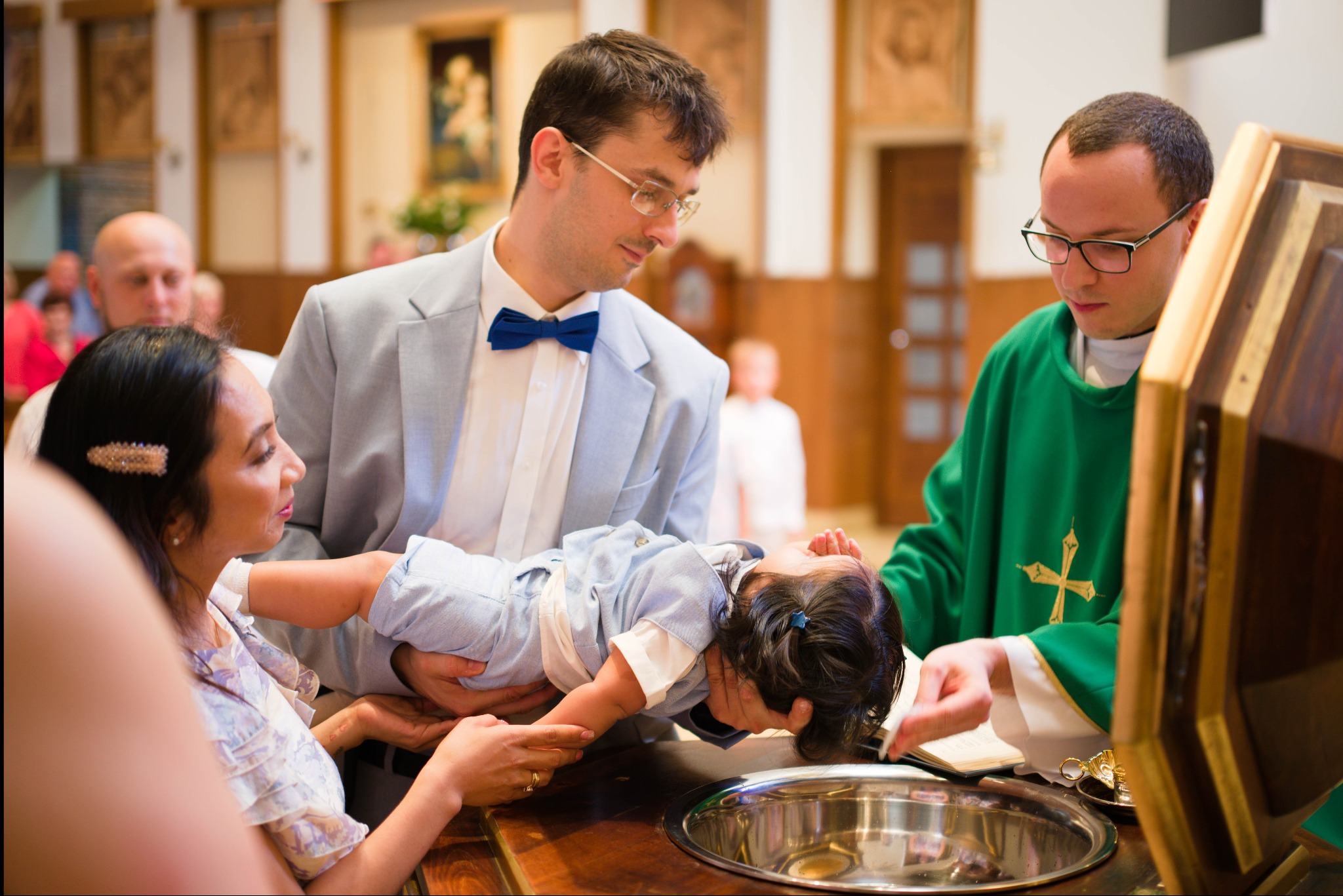 Chrzest dziecka w kościele: ksiądz w zielonej szacie polewa wodą główkę niemowlęcia trzymanego przez rodziców. W tle obraz i inni uczestnicy uroczystości.