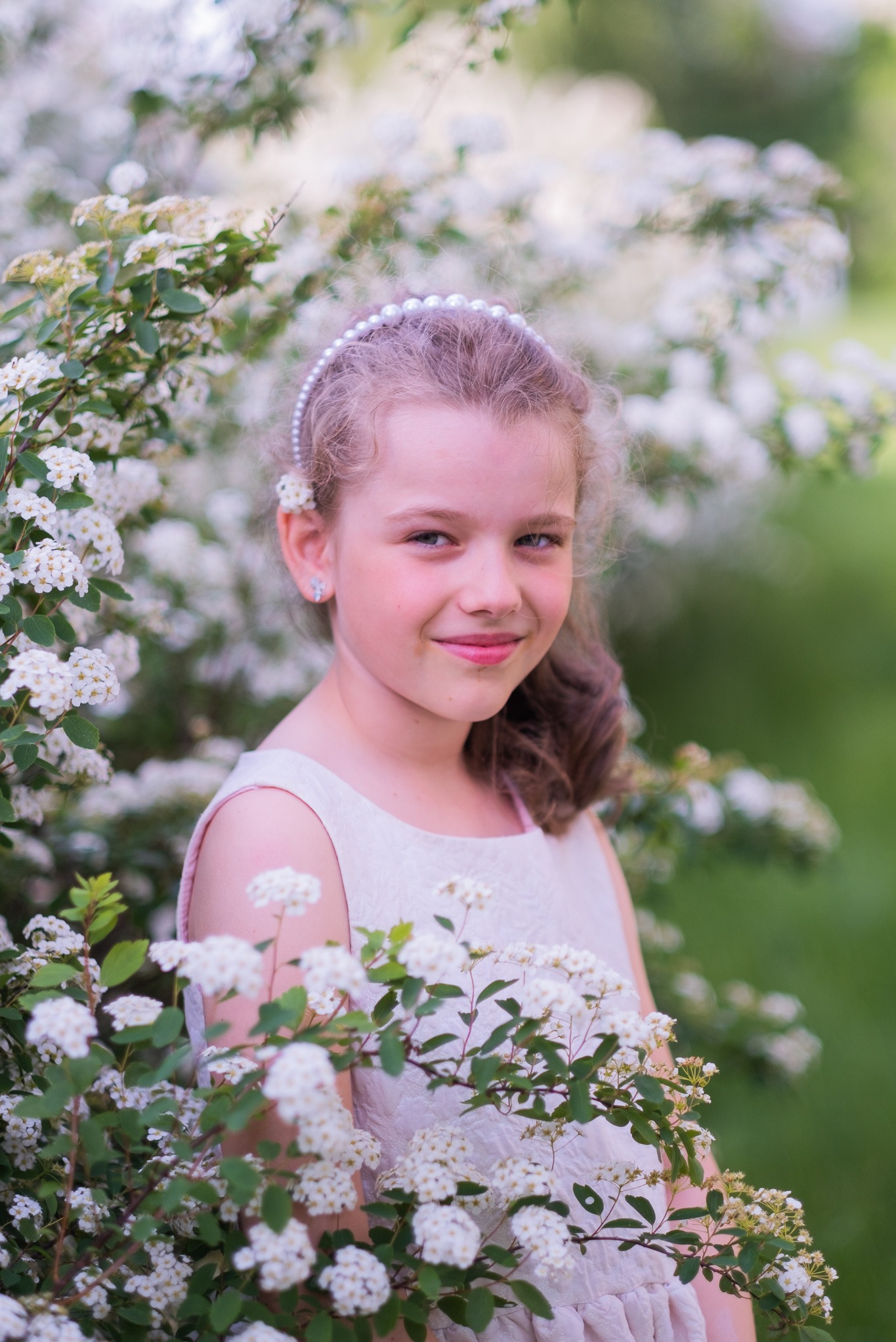 Portret uśmiechniętej dziewczynki w jasnej sukience, otoczonej kwitnącymi gałązkami tawuły. Delikatna opaska z pereł we włosach, naturalne światło.