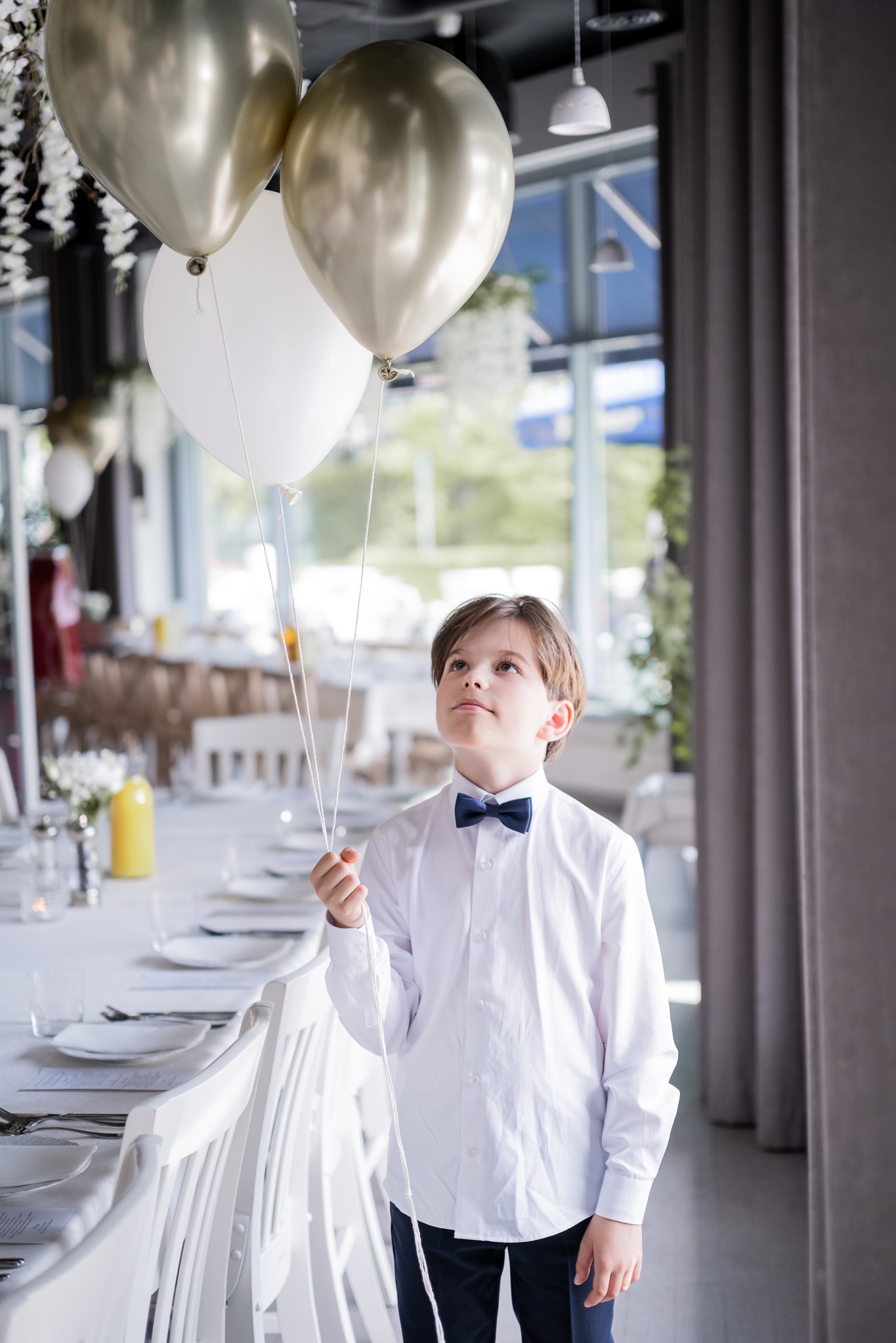 Elegancki chłopiec w białej koszuli i muszce trzymający balony na tle jasnego wnętrza restauracji, uchwycony z dołu, z widocznymi detalami dekoracji stołu.