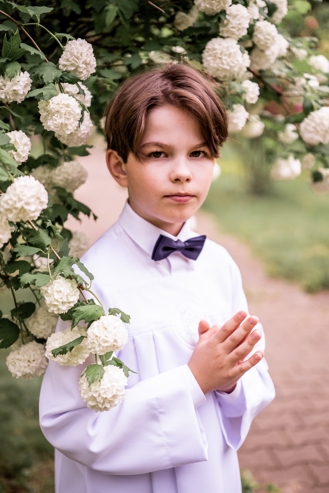 Chłopiec w białej albie i muszce, pozuje na tle kwitnącego krzewu. Ujęcie portretowe, skupione na twarzy i stroju komunijnym.