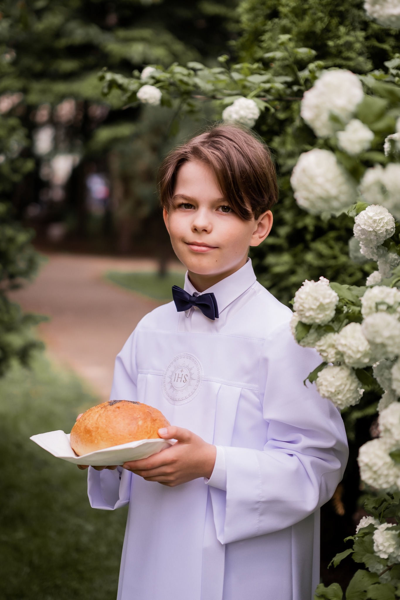 Chłopiec w albie komunijnej z hostią na tle kwitnącego krzewu. Portret z uroczystości Pierwszej Komunii Świętej, naturalne światło, delikatne tło.