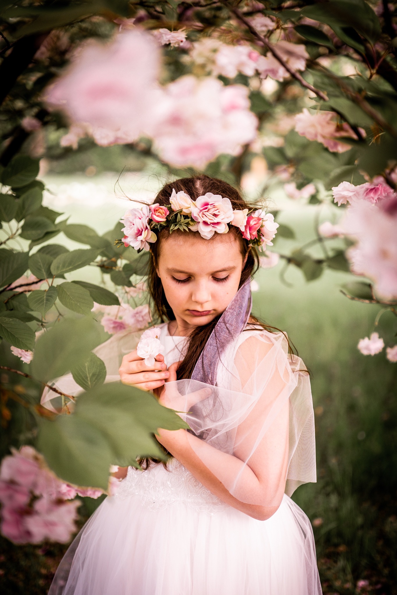 Dziewczynka w białej sukience i wianku z kwiatów, trzymająca piórko na tle kwitnących drzew. Delikatne, naturalne światło podkreśla subtelność sceny.
