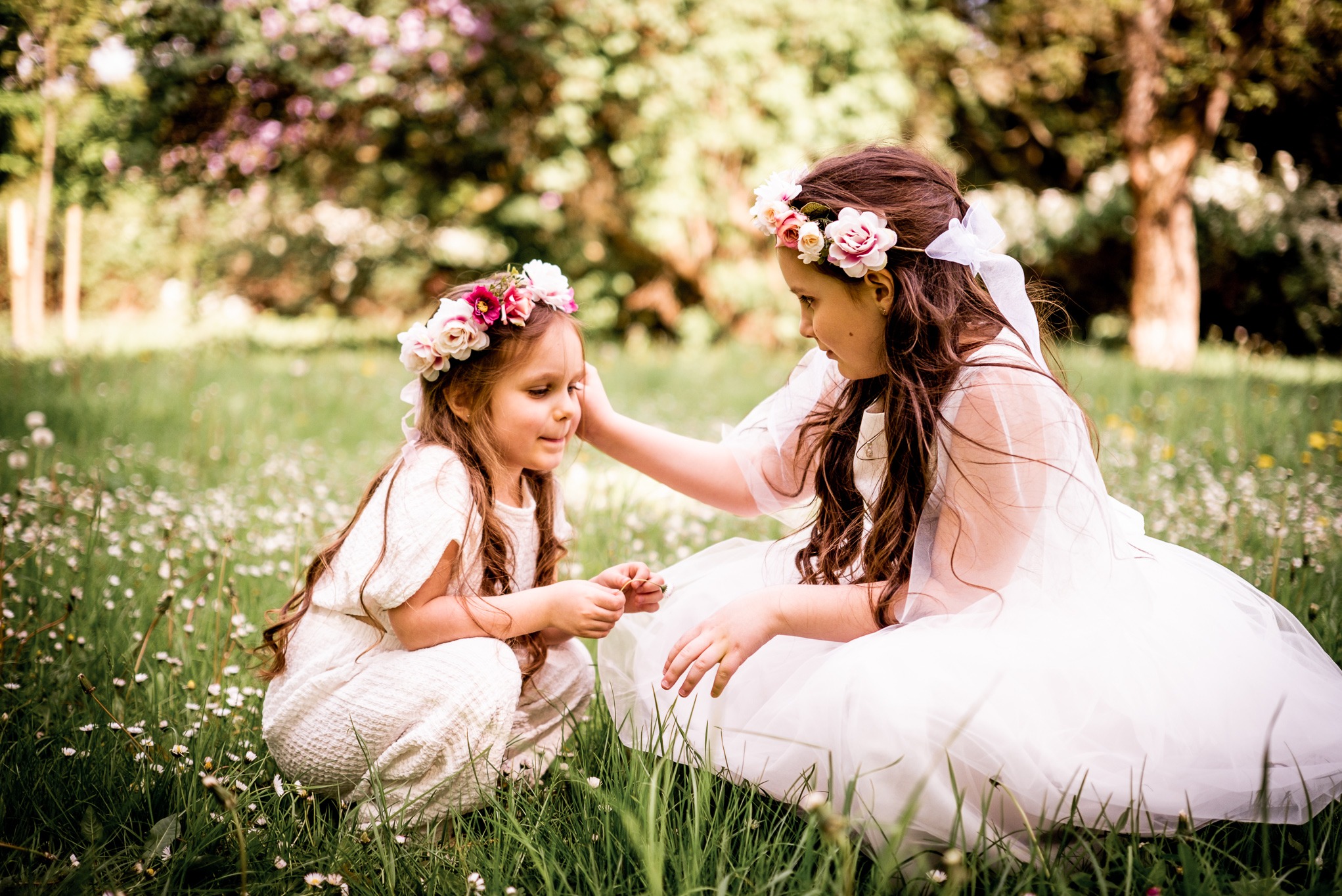Dwie dziewczynki w białych sukienkach i wiankach na głowach siedzą na łące, w tle drzewa. Jedna dotyka włosów drugiej. Delikatne, naturalne światło.