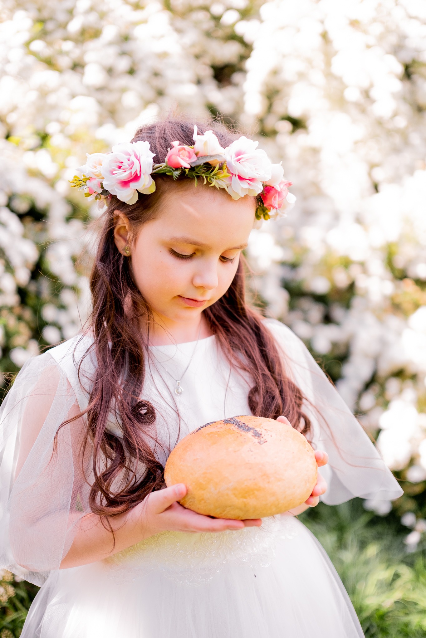 Dziewczynka w białej sukience i wianku z kwiatów trzyma okrągły bochenek chleba na tle kwitnących drzew. Delikatne, rozproszone światło.