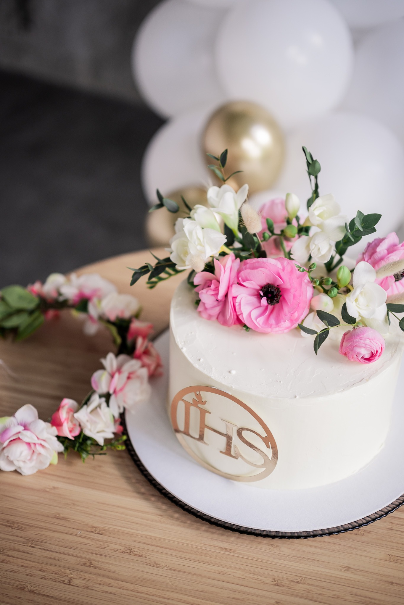 Tort komunijny z białym kremem, złotym symbolem IHS i kwiatową dekoracją na blacie drewnianym, z białymi balonami w tle. Delikatna kompozycja i jasne tło.