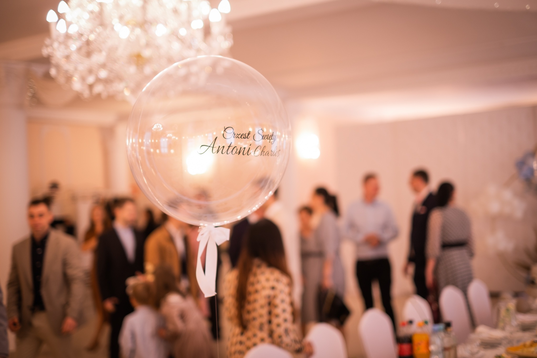 Przezroczysty balon z napisem 'Chrzest Święty Antoni Charles' na tle sali z gośćmi i kryształowym żyrandolem. Dekoracja przyjęcia.
