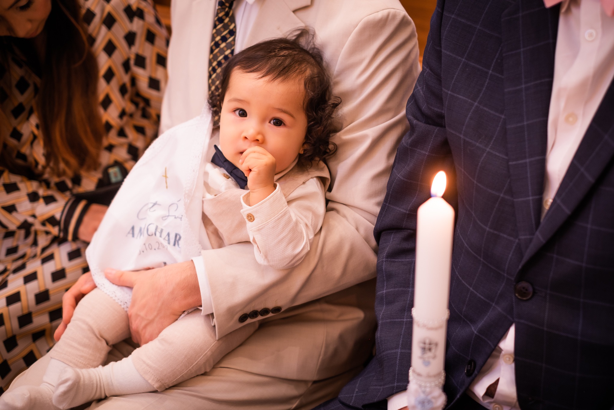 Urocze dziecko w beżowym ubranku i muszce, trzymane na rękach, z zapaloną świecą obok. Pamiątka z Chrztu Świętego z wyhaftowanym imieniem i datą.