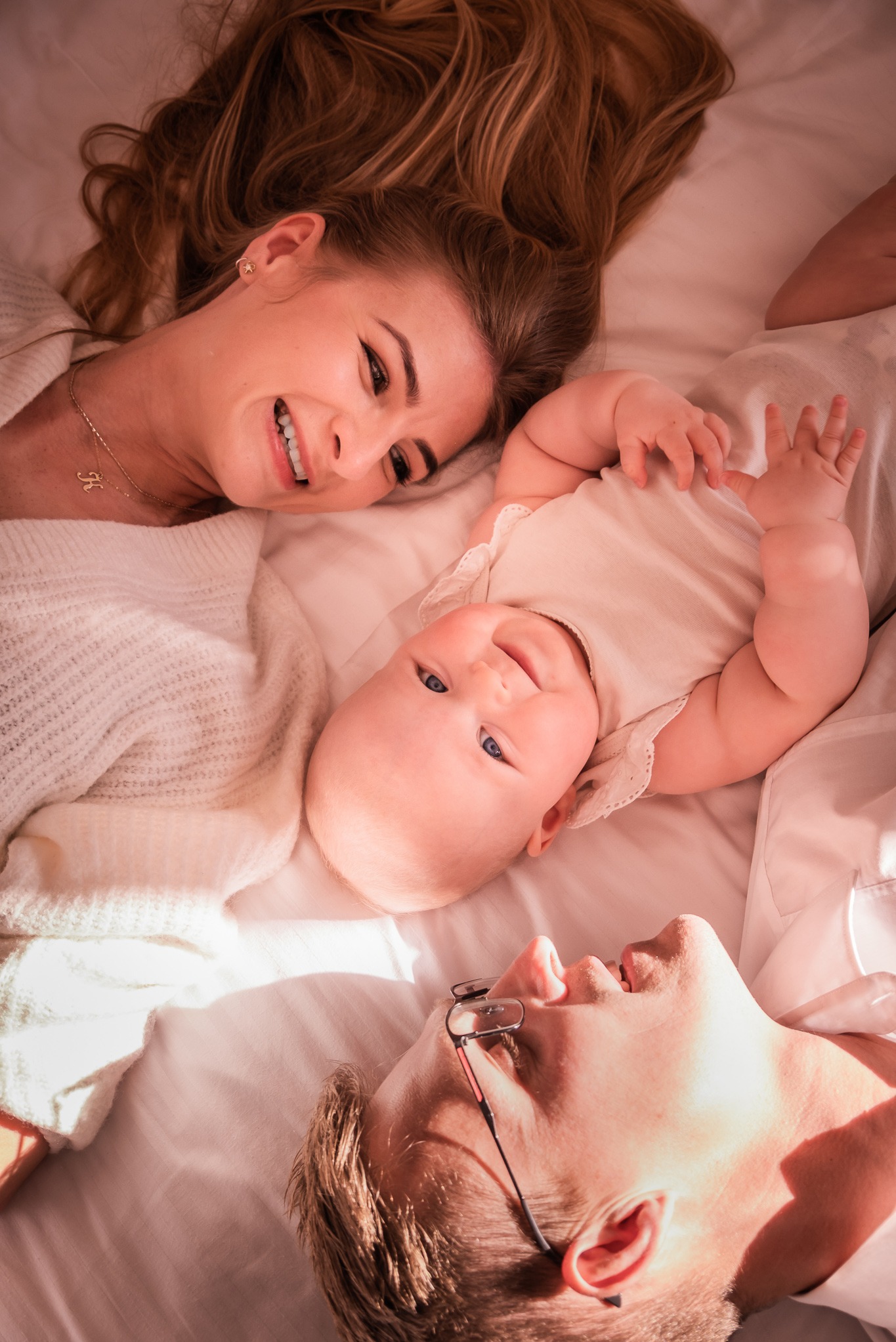 Uśmiechnięta mama, tata w okularach i niemowlę leżą na białym łóżku, patrząc na siebie. Ciepłe, naturalne światło podkreśla bliskość rodziny.