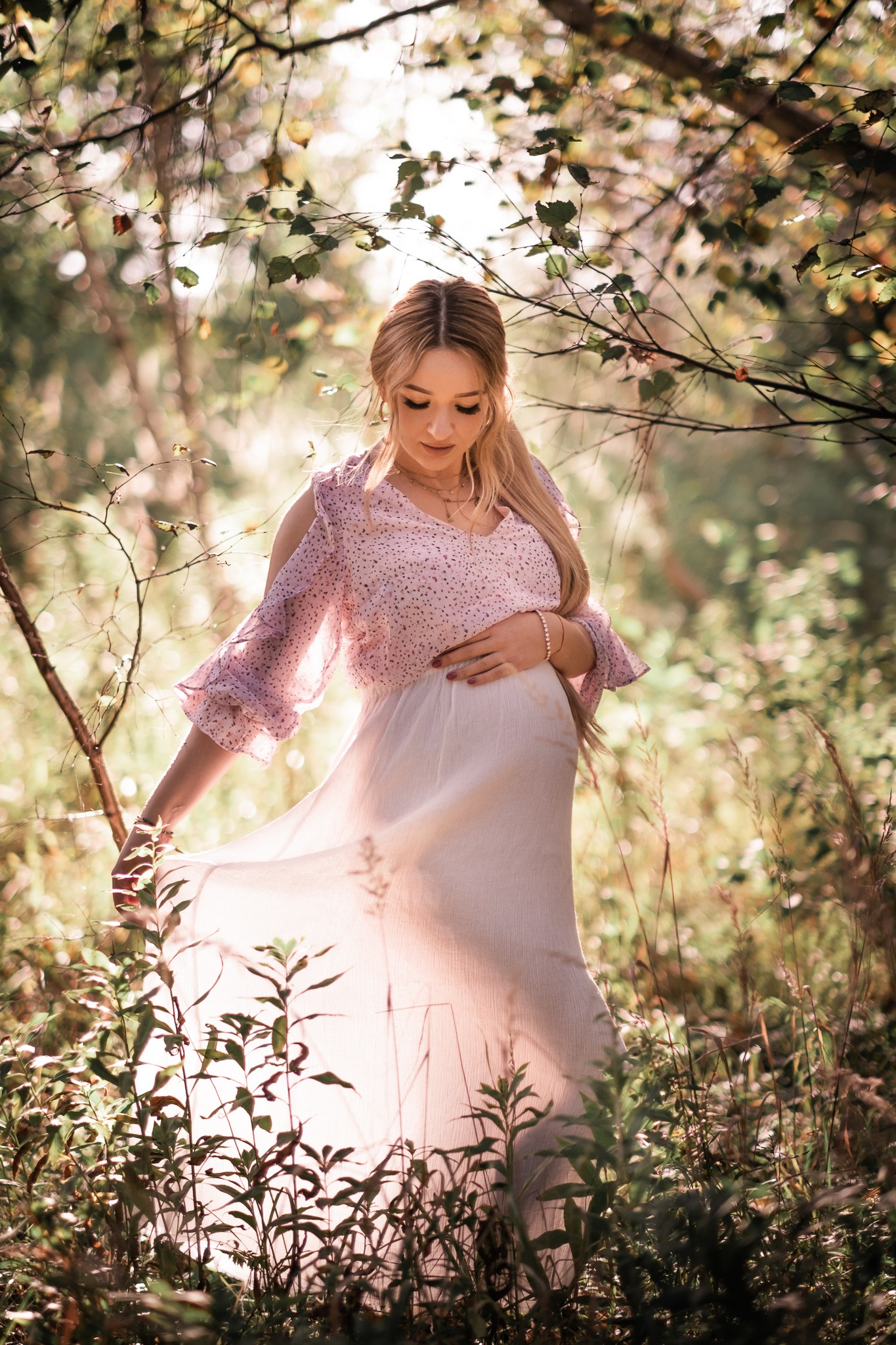 Kobieta w ciąży w zwiewnej sukience, delikatnie dotyka brzucha, pozując w leśnej scenerii z promieniami słońca przebijającymi przez gałęzie. Romantyczna atmosfera.
