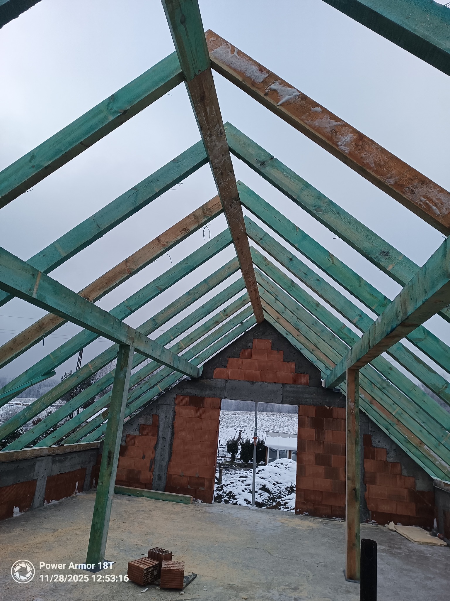 Surowa konstrukcja dachu z pomalowanymi na zielono belkami, widok z wnętrza budynku na zimowy krajobraz. Częściowo wymurowane ściany z czerwonej cegły.