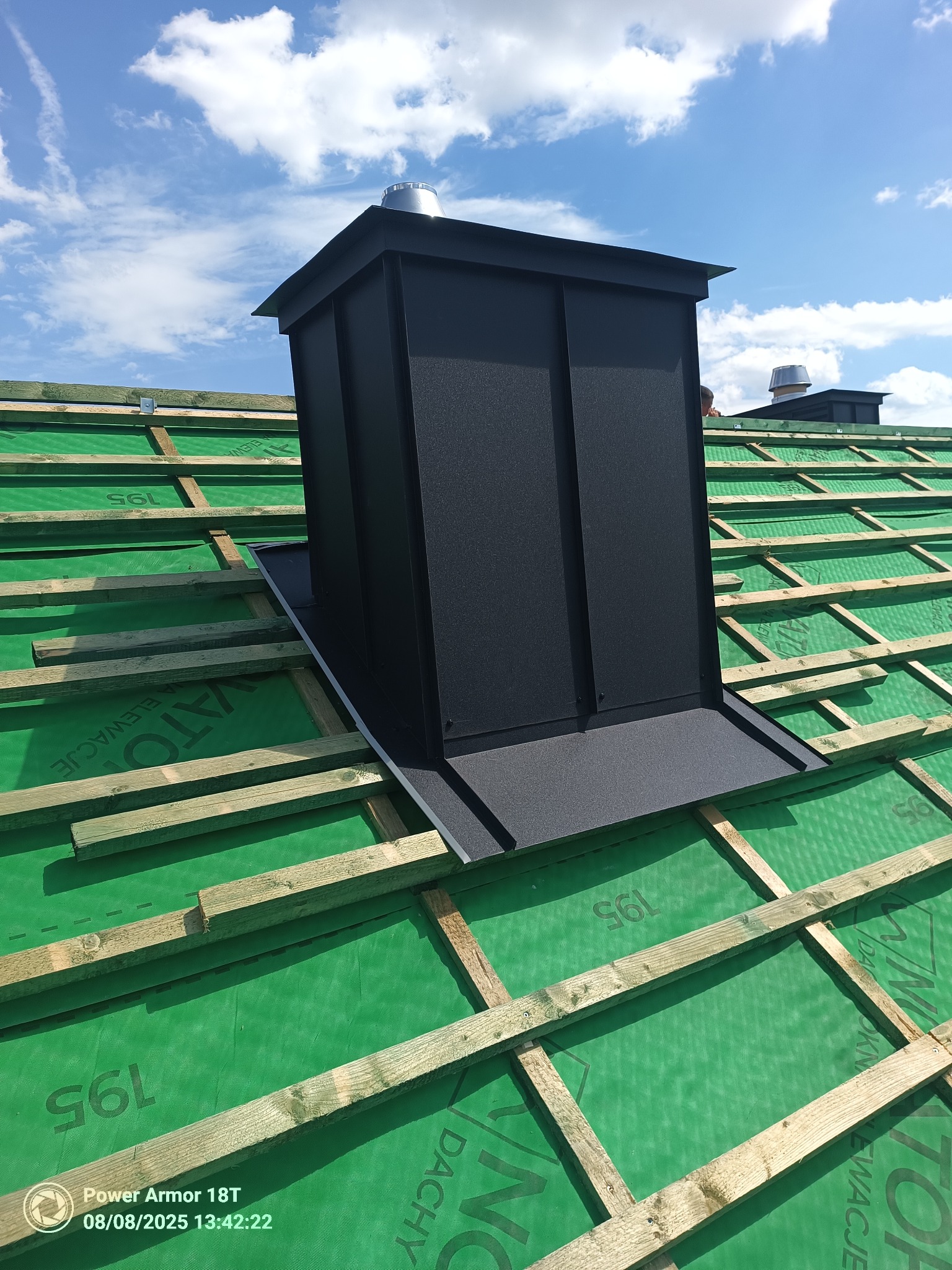 Czarny komin wentylacyjny na dachu z zieloną membraną i drewnianą łatą, widok z góry w słoneczny dzień. Detal obróbki blacharskiej.