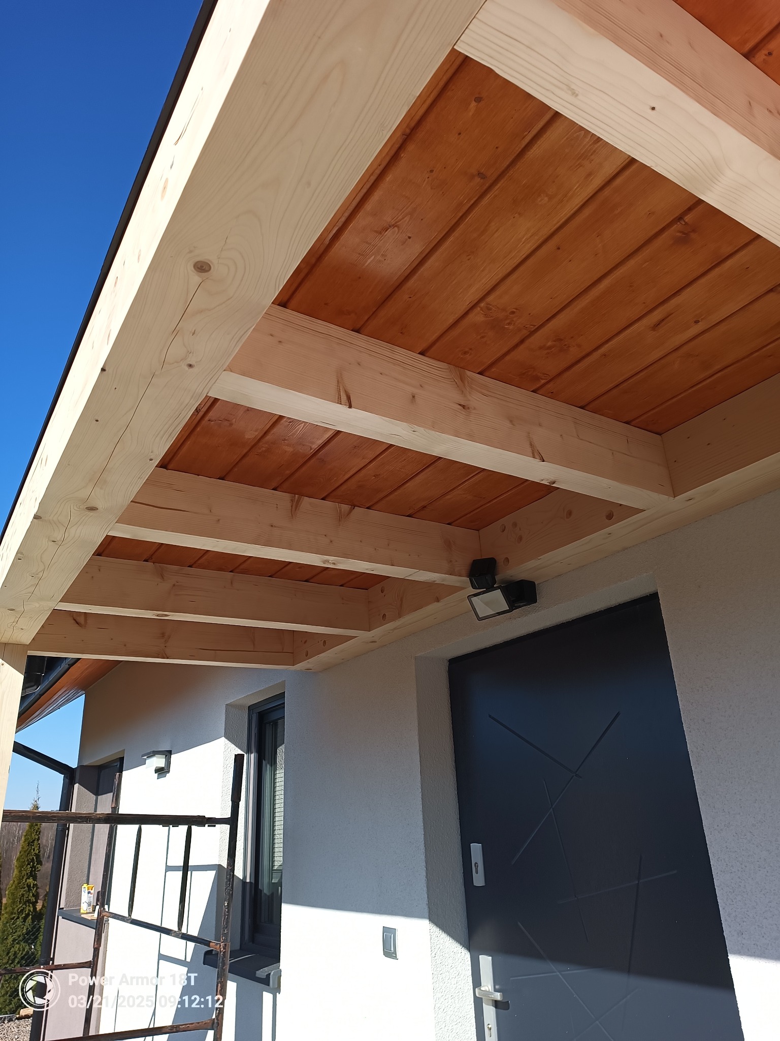 Drewniana konstrukcja z belek i desek, prawdopodobnie zadaszenie tarasu lub werandy, z widocznym oświetleniem zewnętrznym i fragmentem elewacji budynku.