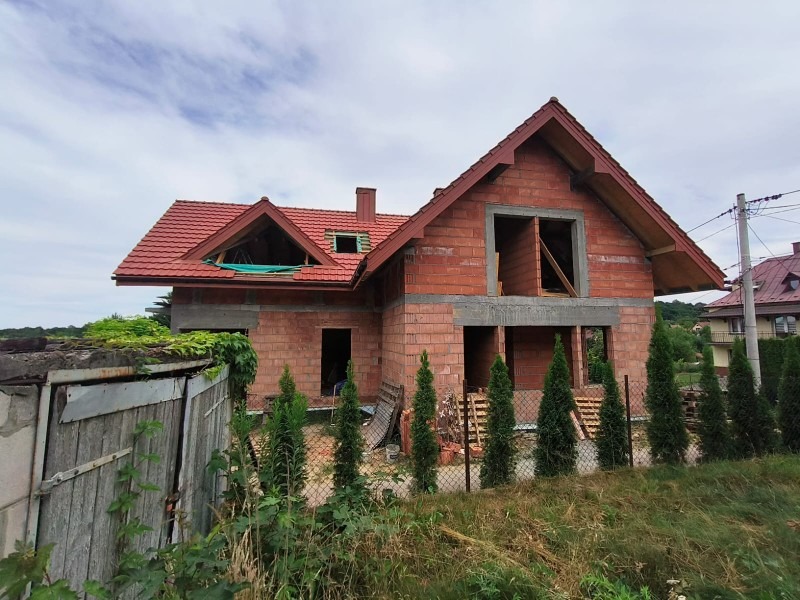 Niedokończony dom z czerwonej cegły z dachem z czerwonej dachówki, widoczne otwory okienne i drzwiowe, w tle zieleń i błękitne niebo.