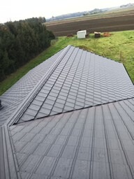 Szary dach z blachy na domu jednorodzinnym, widok z góry, zieleń w tle. Nowoczesne krycie dachu z wyraźną fakturą, równomierne ułożenie blachodachówki.