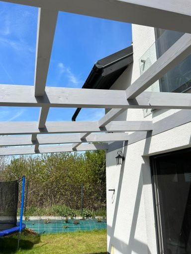 Szara pergola przy białym domu z czarną rynną na tle błękitnego nieba i zieleni ogrodu. Widok z dołu, podkreślający konstrukcję i jej połączenie z budynkiem.