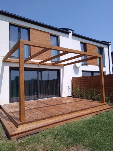 Drewniany taras z pergolą przy nowoczesnym domu w Łętowni. Deski tarasowe ułożone w regularny wzór, widoczne schodki i trawnik. Jasne, naturalne oświetlenie.