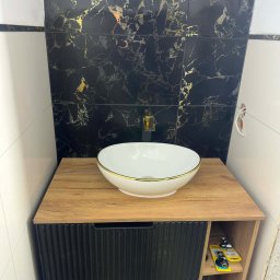 Umywalka nablatowa w złotej obwódce na szafce z czarnymi frontami, na tle ściany z czarno-złotych płytek imitujących marmur. Nowoczesna aranżacja łazienki.