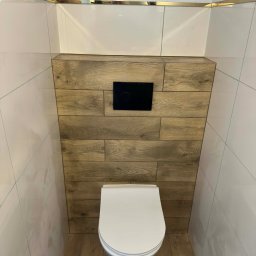 Nowoczesna toaleta podwieszana z drewnianą obudową stelaża i białymi płytkami. Złote listwy dekoracyjne podkreślają elegancki charakter wykończenia.