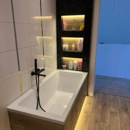 Nowoczesna łazienka z białą wanną obudowaną drewnem, czarną armaturą i podświetlanymi półkami w czarnej, marmurkowej wnęce. Białe płytki z złotymi akcentami.