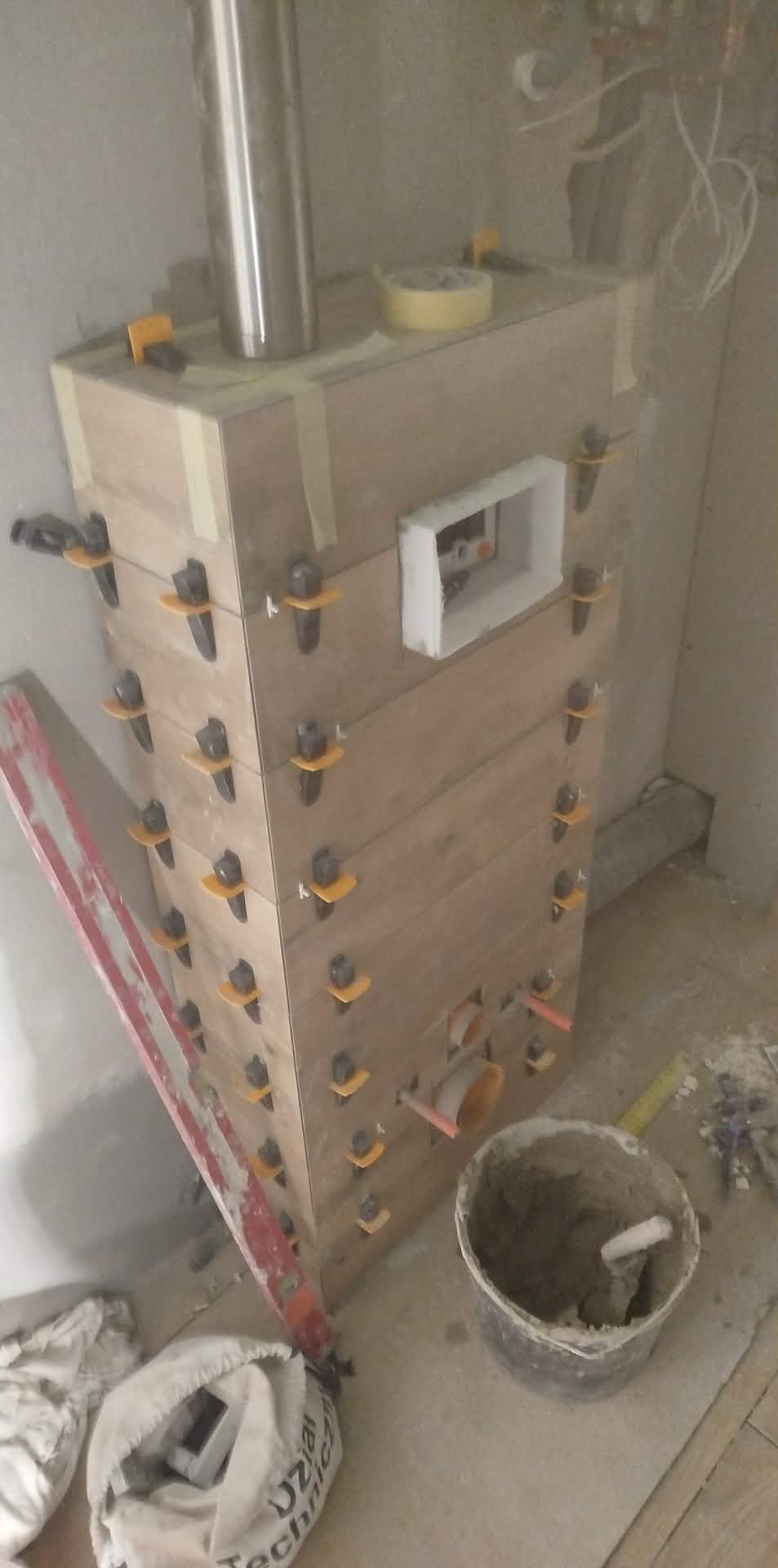 Obudowa z płytek imitujących drewno w trakcie montażu, z użyciem klipsów poziomujących. Widoczny fragment poziomicy i wiadro z zaprawą.