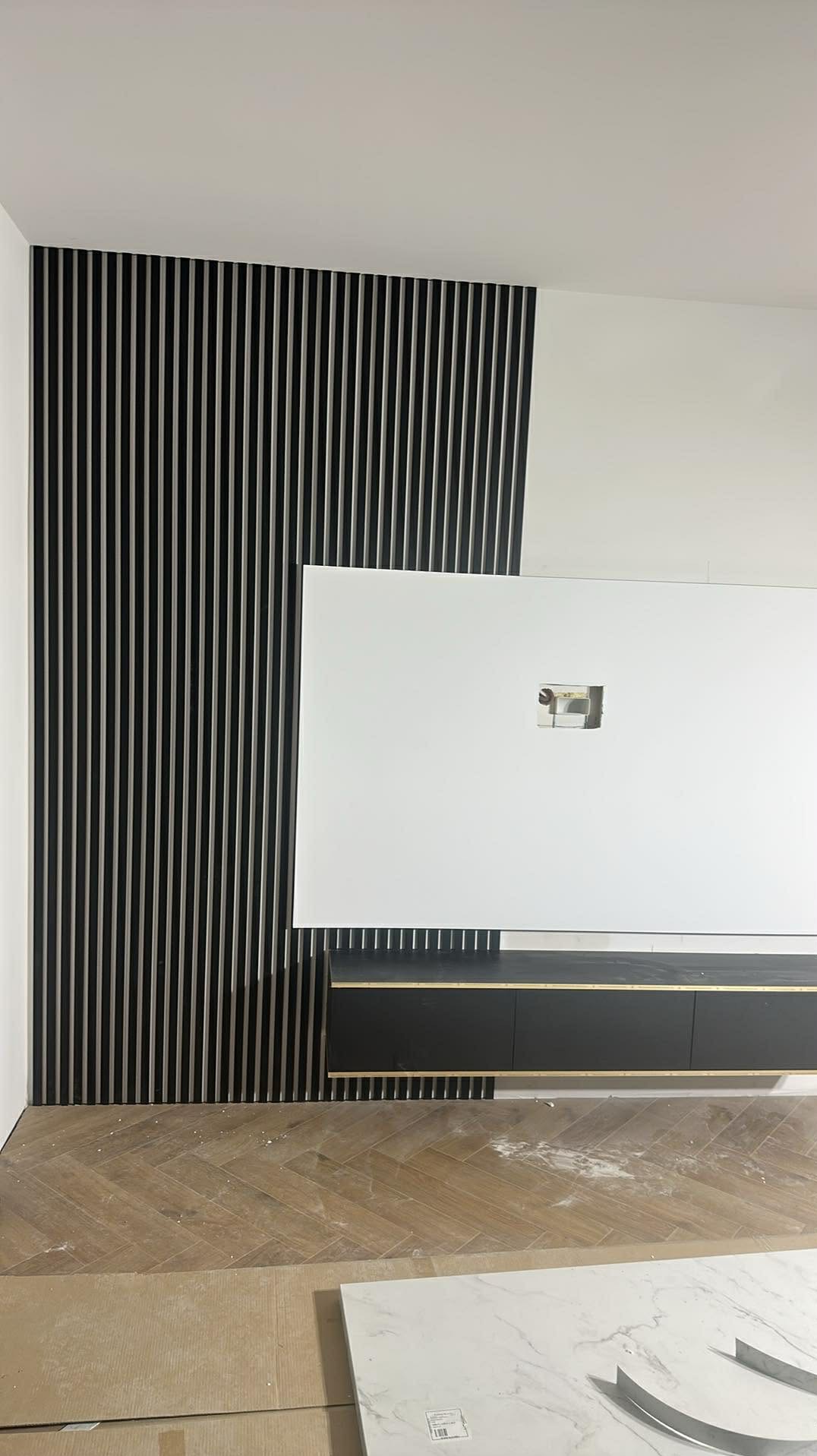 Częściowy widok ściany z dekoracyjnymi, pionowymi panelami w kolorze czarnym, białej płyty i czarnej szafki. Na podłodze panele ułożone w jodełkę.