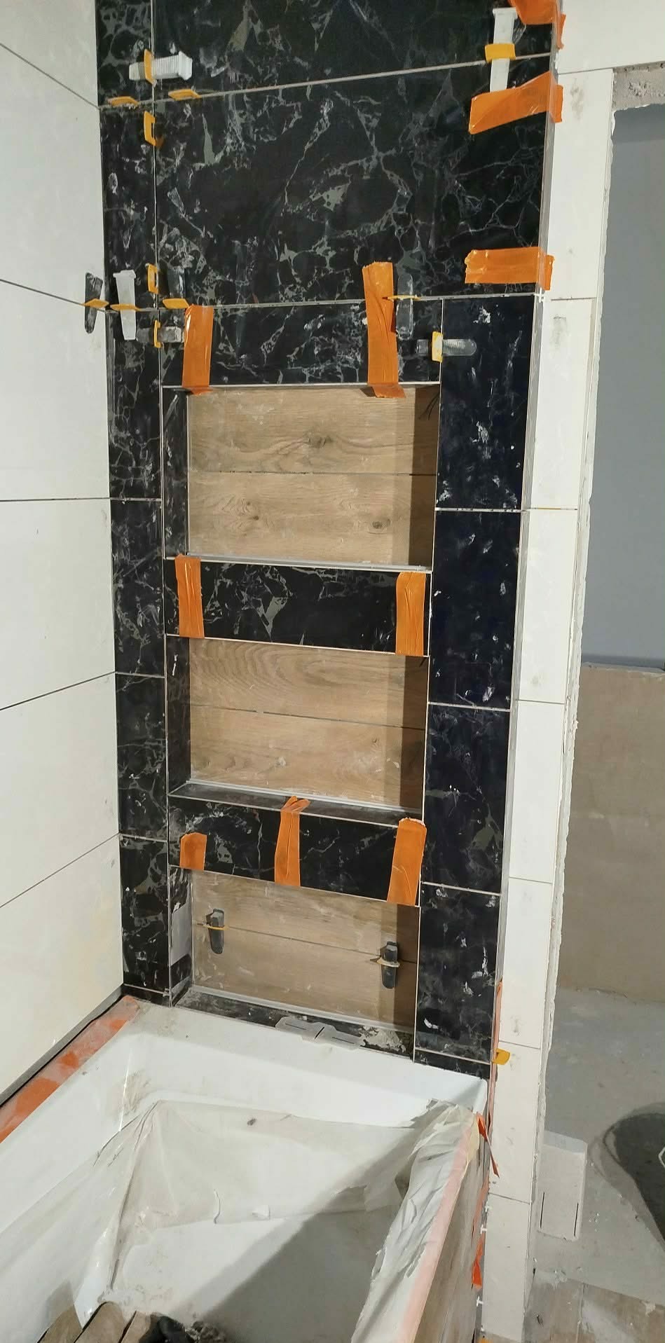 Montaż płytek w łazience: czarne marmurowe płytki z drewnianymi wnękami, pomarańczowa taśma maskująca i klipsy poziomujące, widok na wannę zabezpieczoną folią.