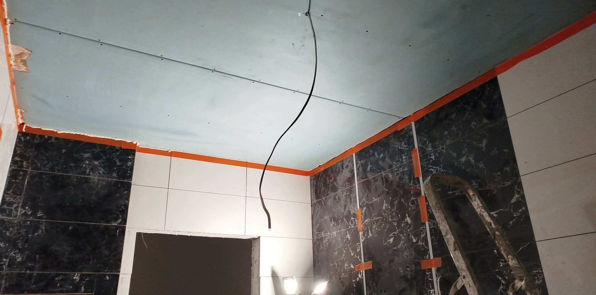 Wnętrze łazienki w trakcie wykańczania: położone płytki imitujące marmur i białe, podwieszany sufit z wystającym kablem, zabezpieczone krawędzie, drabina.