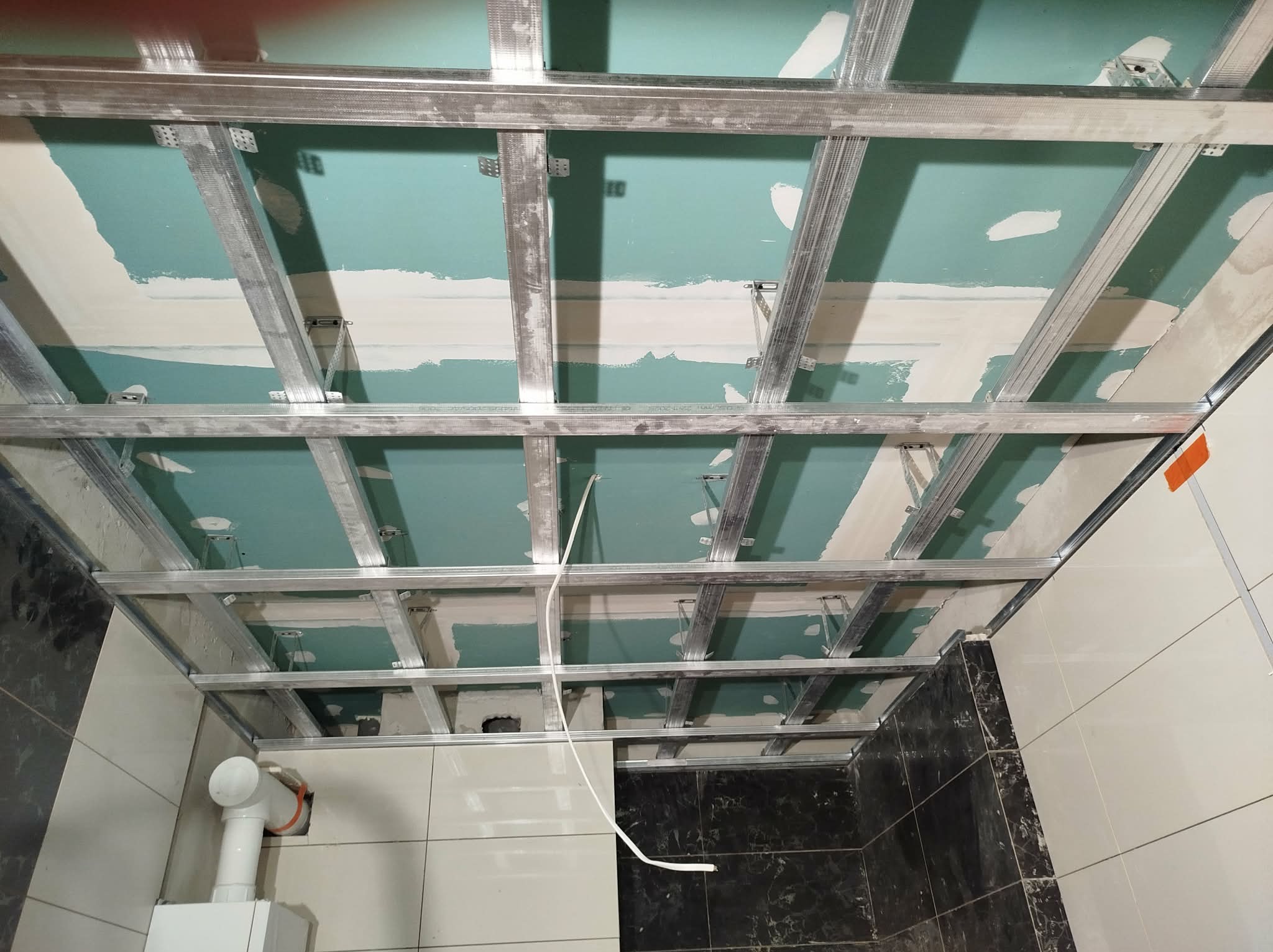 Sufit podwieszany z metalową konstrukcją i płytami gipsowo-kartonowymi w trakcie montażu w łazience z częściowo wykończonymi ścianami z płytek.