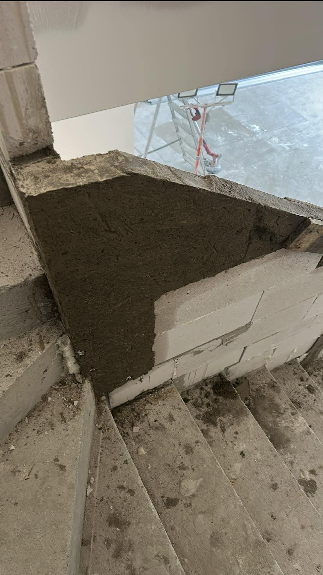 Wnętrze w trakcie prac murarskich: fragment ściany z betonu komórkowego, częściowo pokrytej szarą zaprawą, widoczne schody i rusztowanie w tle.