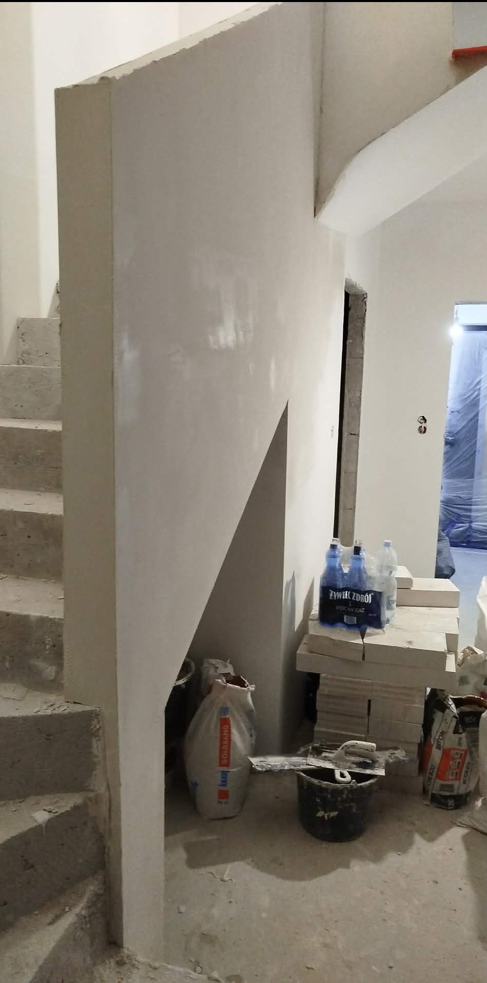 Świeżo wyszpachlowana ściana przy schodach, widoczne narzędzia i materiały budowlane. W tle drzwi zabezpieczone folią malarską.