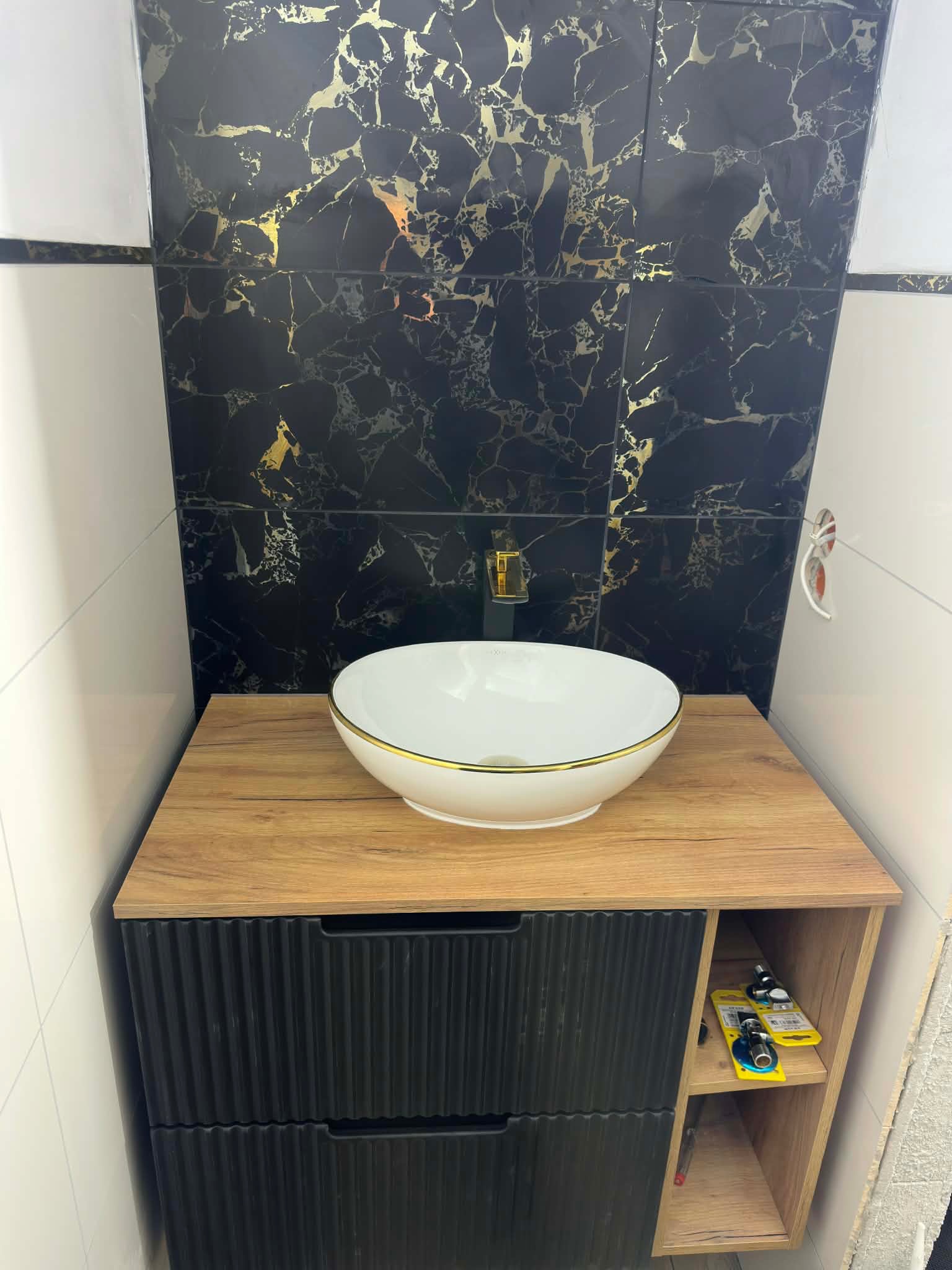 Umywalka nablatowa w złotej obwódce na szafce z czarnymi frontami, na tle ściany z czarno-złotych płytek imitujących marmur. Nowoczesna aranżacja łazienki.