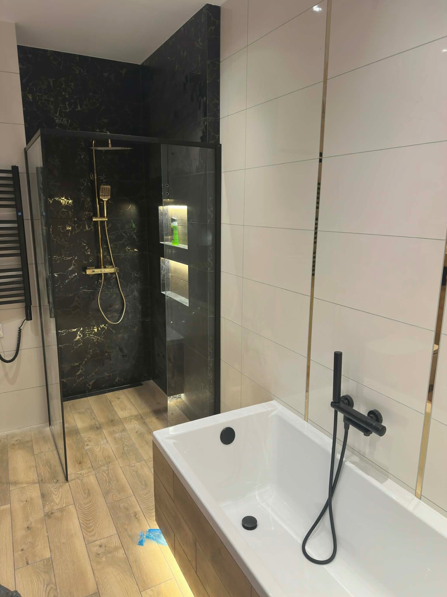 Nowoczesna łazienka z kabiną prysznicową typu walk-in, czarnymi płytkami imitującymi marmur ze złotymi żyłami i białą wanną z czarną armaturą.
