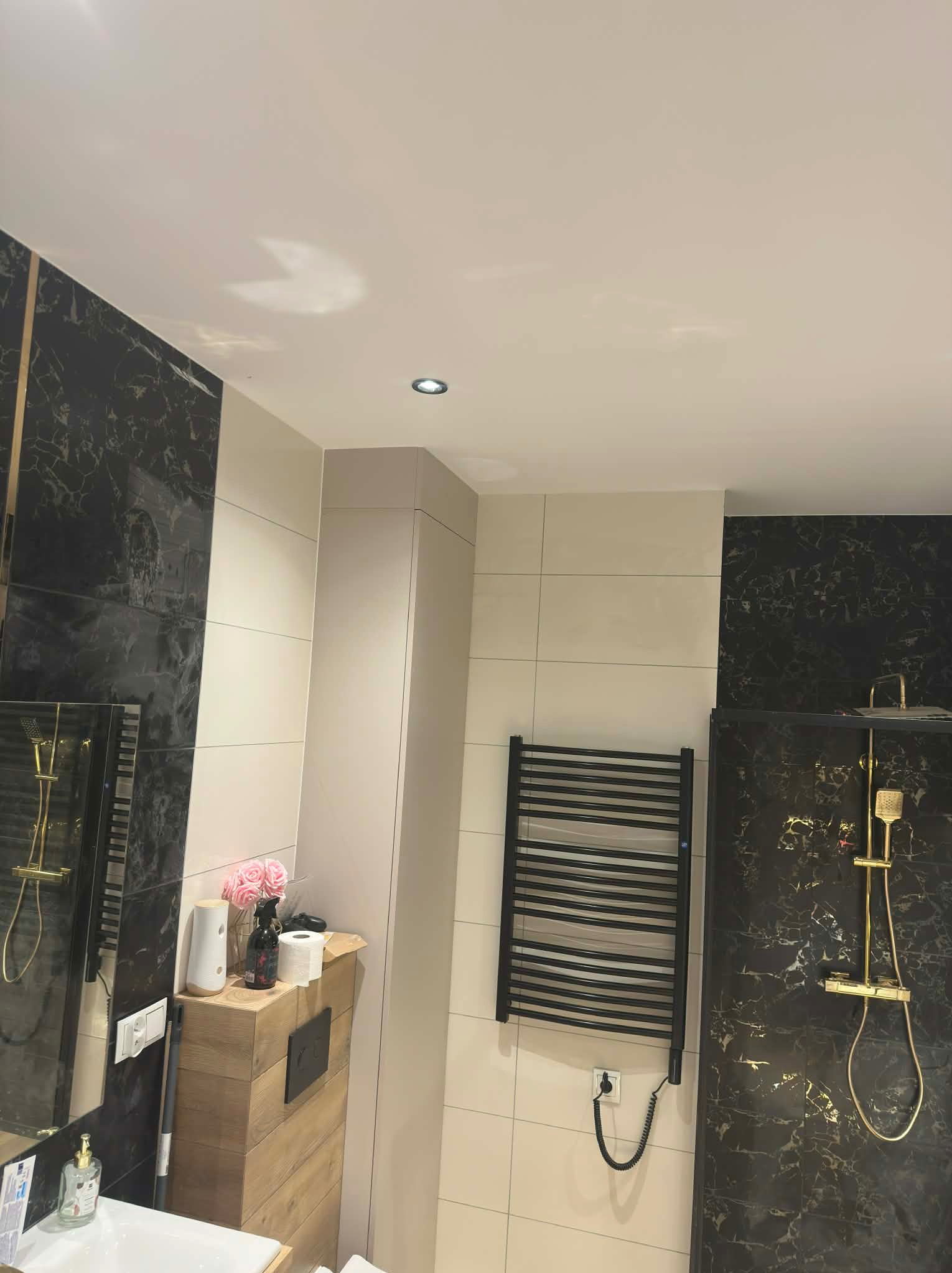 Elegancka łazienka po remoncie: czarne marmurowe płytki, beżowe ściany, drewniana szafka z kwiatami i czarny grzejnik drabinkowy. Nowoczesny design z dbałością o detale.