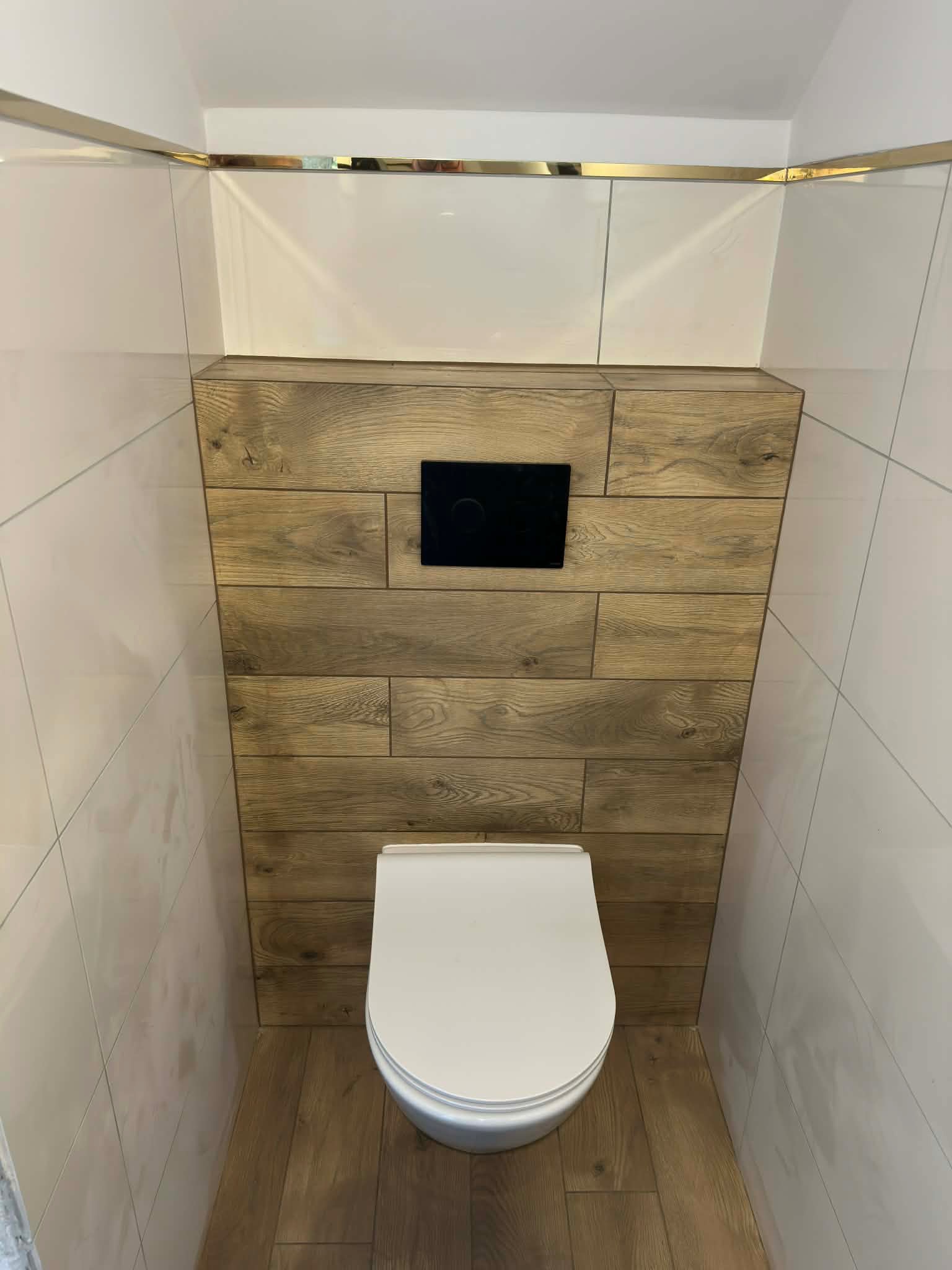 Nowoczesna toaleta podwieszana z drewnianą obudową stelaża i białymi płytkami. Złote listwy dekoracyjne podkreślają elegancki charakter wykończenia.