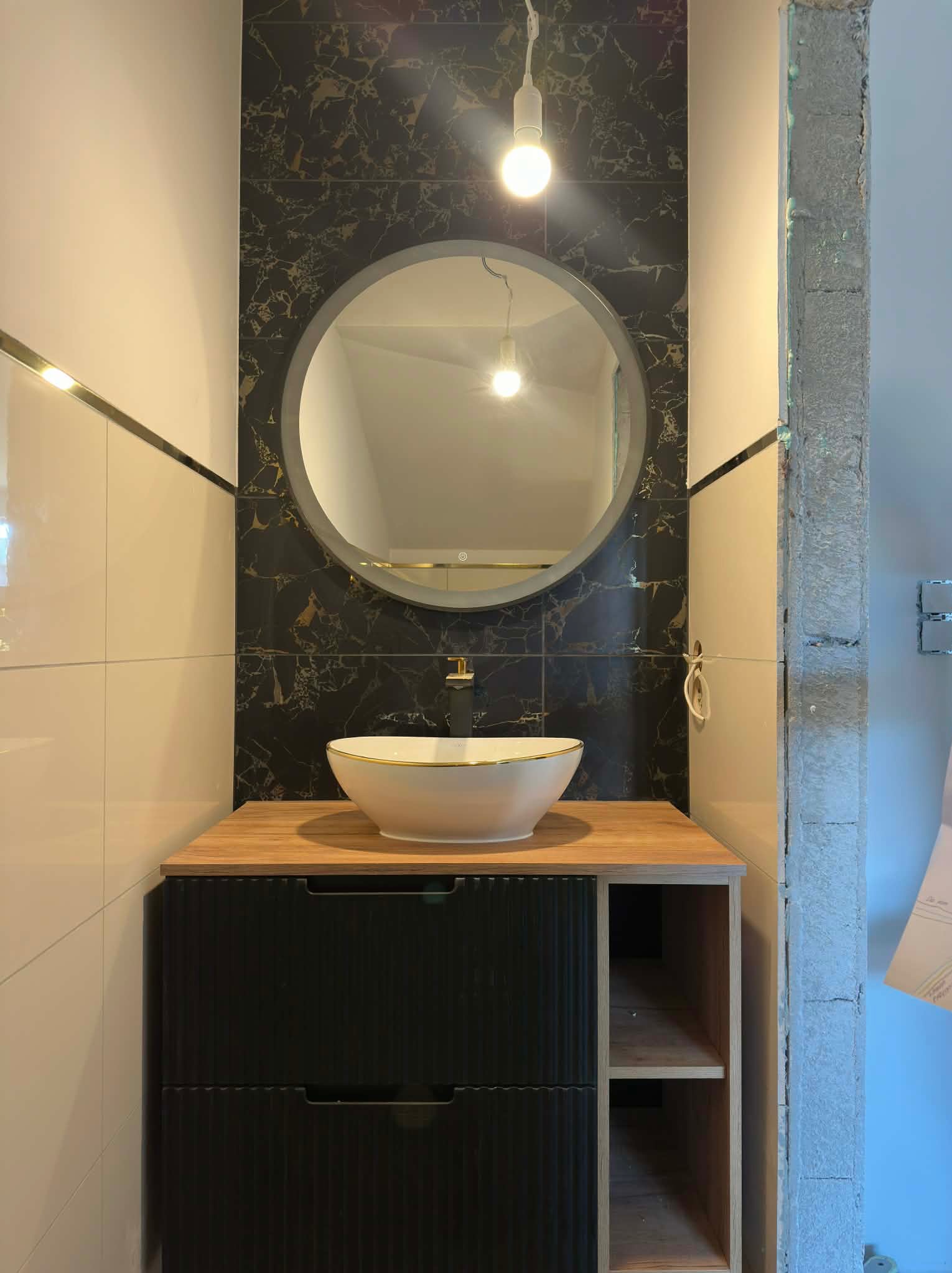 Nowoczesna łazienka po remoncie: okrągłe lustro z podświetleniem nad białą umywalką na szafce z czarnymi frontami. Marmurowe płytki i oświetlenie punktowe.
