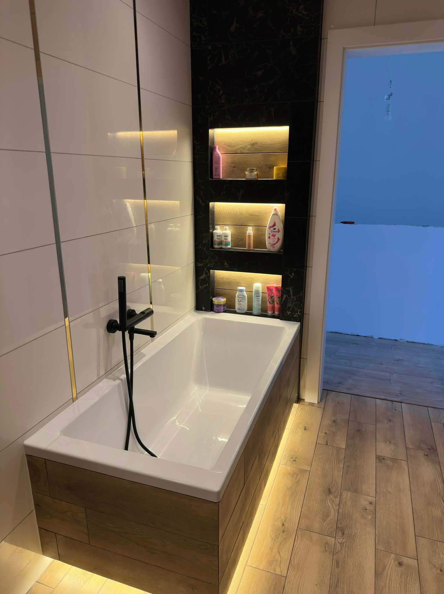 Nowoczesna łazienka z białą wanną obudowaną drewnem, czarną armaturą i podświetlanymi półkami w czarnej, marmurkowej wnęce. Białe płytki z złotymi akcentami.