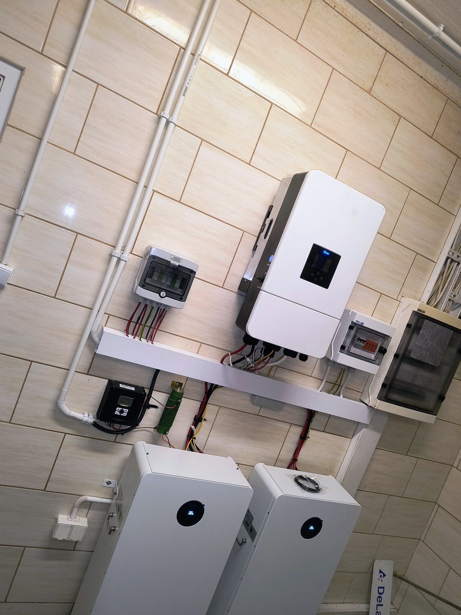 Kompletna instalacja fotowoltaiczna w pomieszczeniu: biały inwerter, akumulatory, rozdzielnice i okablowanie na tle beżowych płytek ściennych.