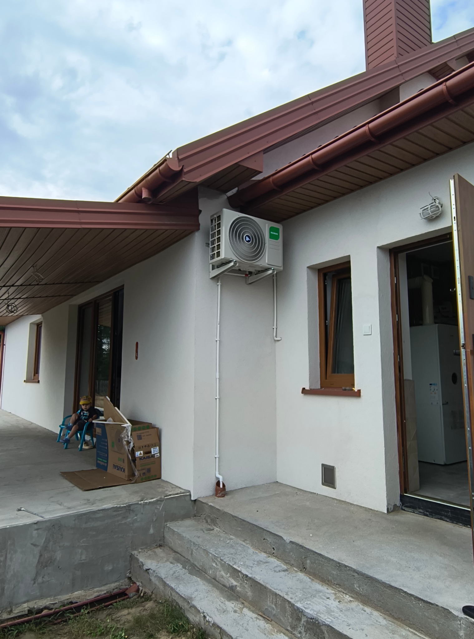Biały klimatyzator Kaisai zamontowany na ścianie domu z brązowym dachem i oknami. Na werandzie siedzi dziecko obok kartonów po klimatyzacji.