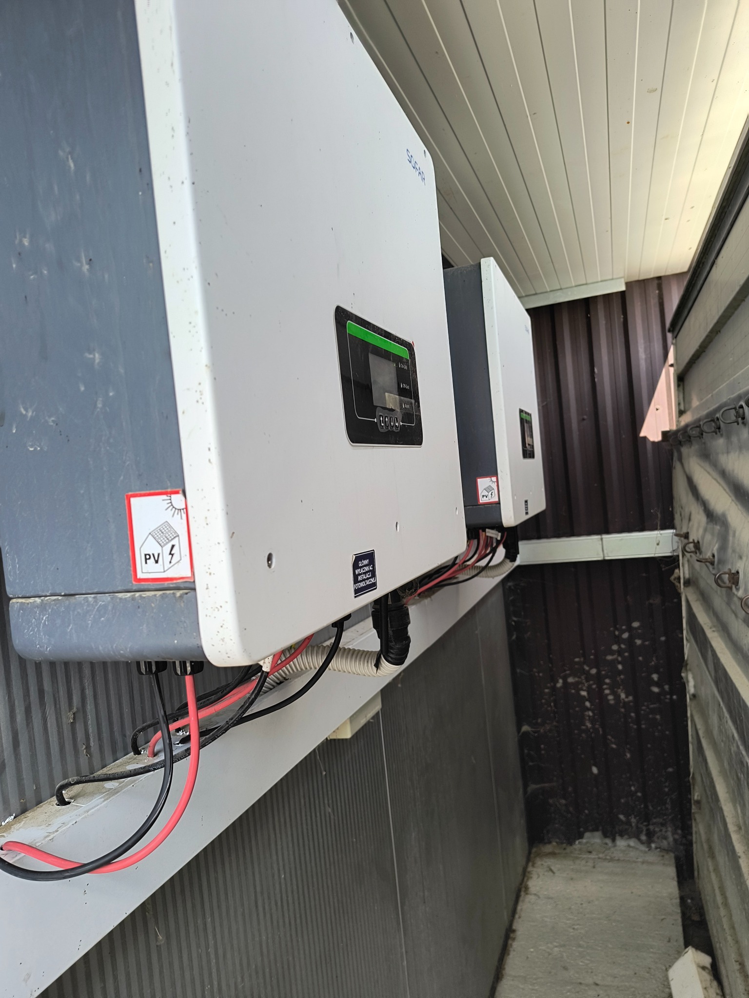 Dwa białe falowniki fotowoltaiczne marki Sofar zamontowane na ścianie z widocznymi kablami i naklejką PV, z perspektywy lekko od dołu.